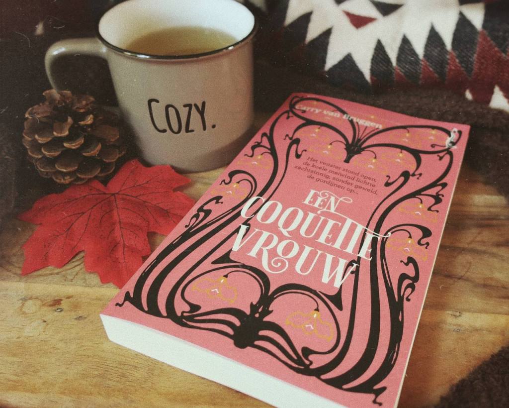 Een coquette vrouw ligt op een houten onderzetter. Daarnaast liggen herfstdecoraties en staat een grijze mok met daarop het woord 'Cozy.' 