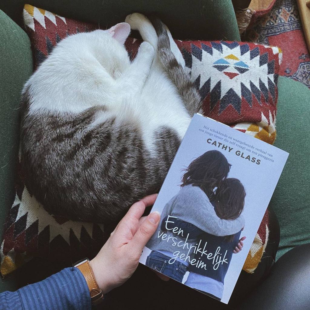 Ik houd het boek Een verschrikkelijk geheim van Cathy Glass vast op de bank. Naast het boek ligt Alfie mijn grijs-witte kat te slapen op een rood kussen.