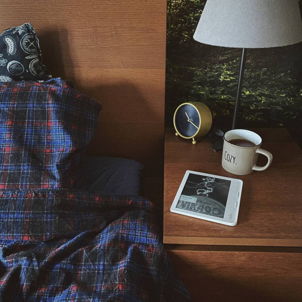 Een deel van mijn bed met daarnaast mijn nachtkastje. Daarbovenop ligt mijn e-reader met Virago erop open, er staat een grote mok thee naast met het woord 'Cozy.' erop. 