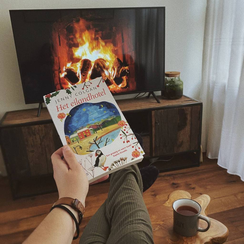 Een gezellig tafereel met een kop warme thee, het boek het Eilandhotel van Jenny Colgan en een knapperend haardvuur op de televisie
