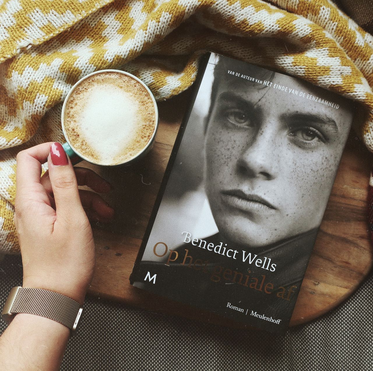 Slider Benedict Wells Op het geniale af recensie Benedict Wells Op het geniale af ligt op een houten ondergrond. Daarnaast staat een kopje koffie die ik met mijn linkerhand vasthoud. Mijn nagels zijn in een roodroze kleur gelakt.