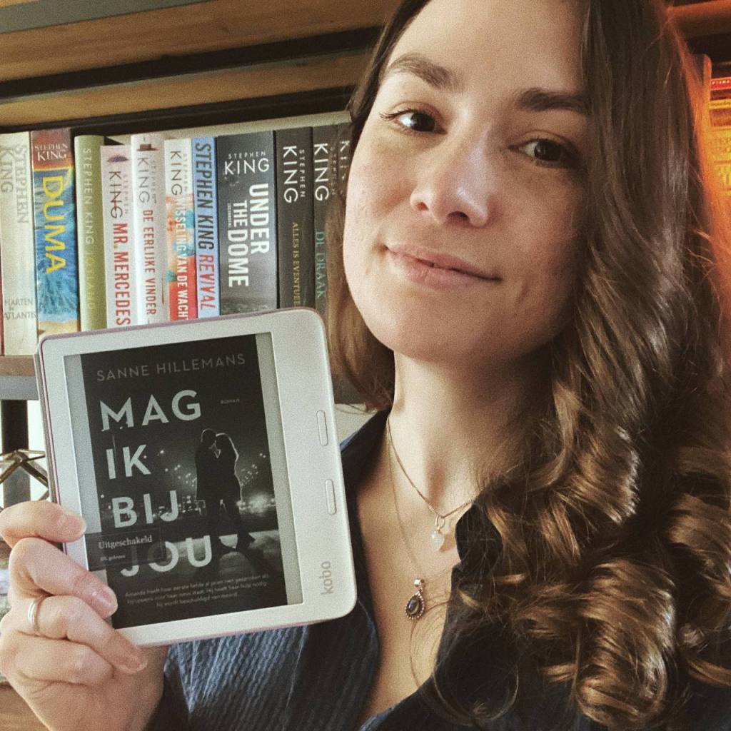 Een selfie. Ik houd voor mijn boekenkast mijn e-reader met daarop het omslag van Mag ik bij jou van Sanne Hillemans vast.