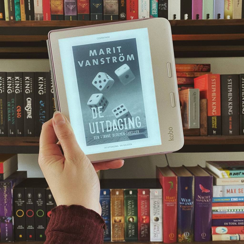 Ik houd mijn Kobo-ereader met daarop het ebook van De uitdaging vast voor de boekenkast.