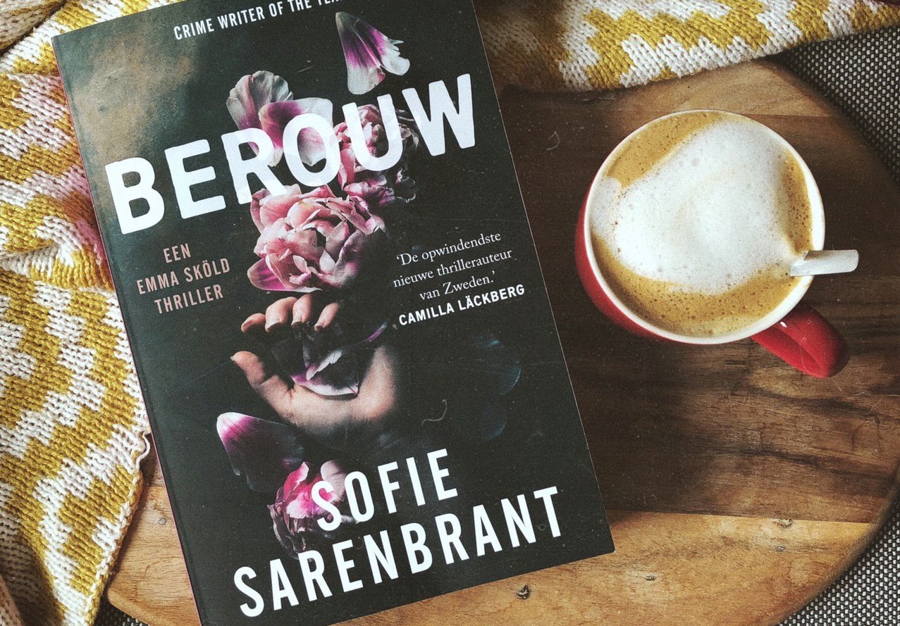 Het boek Berouw van Sofie Sarenbrant ligt op een houten onderlegger. Daarnaast staat een kopje cappuccino en er ligt een dekentje omheen
